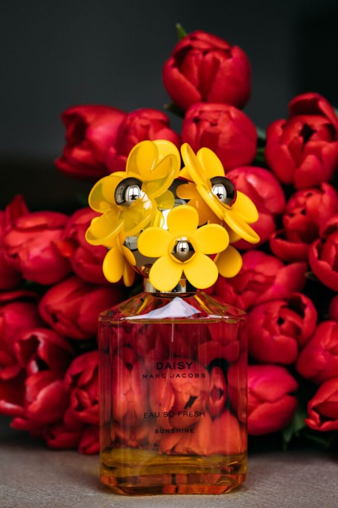 Glass bottle with female fragrance with creative lid in shape of yellow flowers placed on table against wonderful red tulips bouquet
