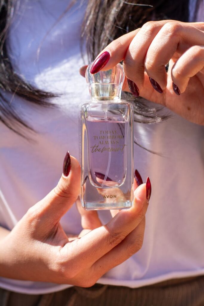 A woman with painted nails holding a clear perfume bottle, captured in natural sunlight.